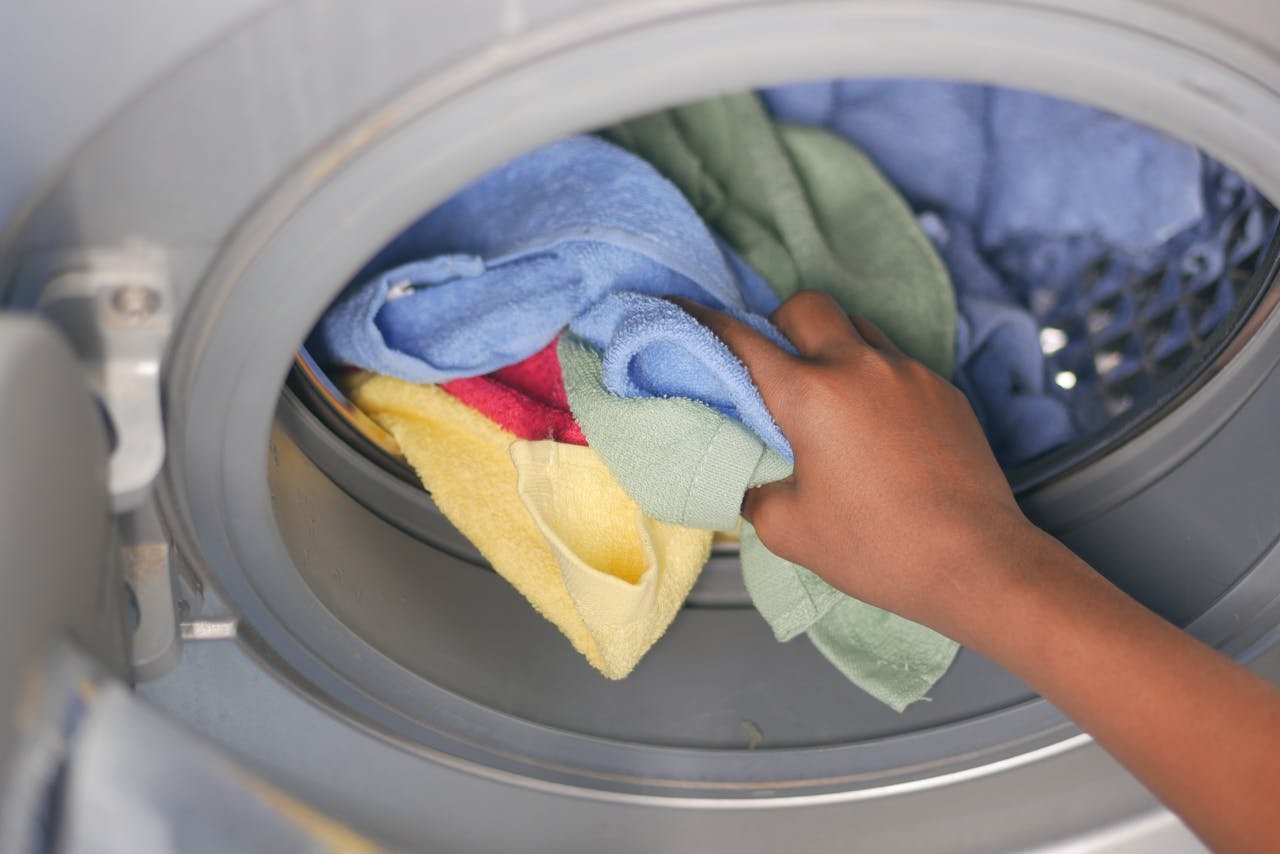 A close-up of a hand loading colorful towels into a front-loading washing machine.